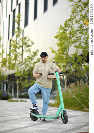 Young courier man using phone, calls client near parked electric scooter, delivery cardboard boxes on street in business area. Concept of sustainability, delivering and eco-friendly modern lifestyle 126133180