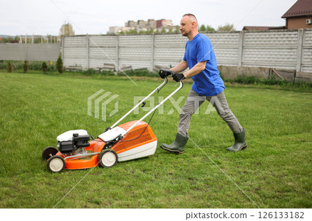 Man in protective gloves, gardener is mowing bright lush green grass lawn using modern gasoline cordless lawnmower at own backyard. Seasonal landscaping design work. Gardening work tools. Man in protective gloves, gardener is mowing bright lush green grass lawn using modern gasoline cordless lawnmower at own backyard. Seasonal landscaping design work. Gardening work tools. 126133182