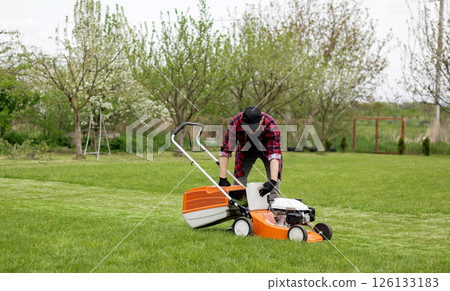 Man in casual clothes takes out bag with cut grass of lawn mower after mowing lawn with at own backyard. Husband takes care of garden. Modern gasoline garden equipment. Landscaping work at home 126133183