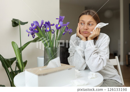 Seasonal allergy. Unhealthy allergic young woman uses paper tissues, suffers from runny nose, sneezes from flowers pollen at kitchen home by table. Girl with flu, itching or cough, allergy rhinitis Seasonal allergy. Unhealthy allergic young woman uses paper tissues, suffers from runny nose, sneezes from flowers pollen at kitchen home by table. Girl with flu, itching or cough, allergy rhinitis 126133198