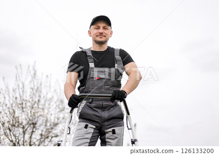 Bottom view of professional male gardener in protective apparel, gloves, cap and overalls holds handle of gasoline cordless lawnmower for mowing grass lawn outdoors. Seasonal landscaping design work Bottom view of professional male gardener in protective apparel, gloves, cap and overalls holds handle of gasoline cordless lawnmower for mowing grass lawn outdoors. Seasonal landscaping design work 126133200