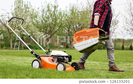 man in casual clothes holds bag with cut grass of lawn mower after mowing lawn with at own backyard. Husband takes care of garden. Modern gasoline garden equipment. Landscaping work at home man in casual clothes holds bag with cut grass of lawn mower after mowing lawn with at own backyard. Husband takes care of garden. Modern gasoline garden equipment. Landscaping work at home 126133201