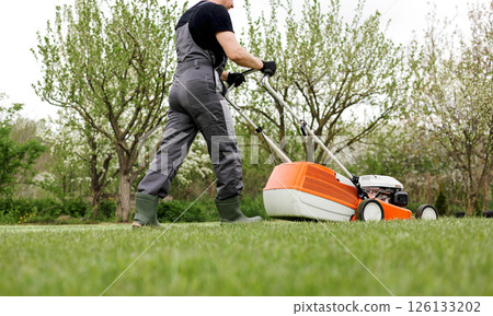 Unrecognizable professional gardener in protective apparel is mowing green grass lawn using modern gasoline cordless lawnmower at backyard. Seasonal landscaping design work. Gardening work tools. Unrecognizable professional gardener in protective apparel is mowing green grass lawn using modern gasoline cordless lawnmower at backyard. Seasonal landscaping design work. Gardening work tools. 126133202