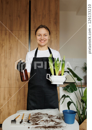 Smiling young florist woman in apron holds hyacinth flowers in new ceramic pot and waters houseplant from water sprayer at home kitchen after repotting. Seasonal work with transplantation plants. 126133210