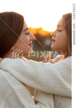 Family day, mother's day. Happy smiling young mom and adorable child daughter soft hugging, kissing and spending time together at autumn on sunset. Idyllic family having fun outdoors on fall holiday. Family day, mother's day. Happy smiling young mom and adorable child daughter soft hugging, kissing and spending time together at autumn on sunset. Idyllic family having fun outdoors on fall holiday. 126133211