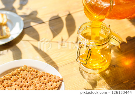 Golden Honey Streams Into a Jar While Light Dances Across the Wooden Table, Accompanied by Crunchy Crackers 126133444