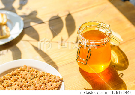 Honey Jar on a Wooden Table With Sunlit Shadows, Crackers, and Pastries, Perfect for Cafes and Bakeries 126133445
