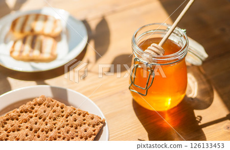 Honey Jar on a Wooden Table With Crisp Bread, Sunlight Casting Shadows ideal for Culinary Blogs and Food Styling 126133453