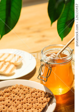 Honey Jar and Wooden Dipper Next to Crispbread and Cookies on a Sunny Wooden Table, Evoking Cafes and Wellness Food 126133457