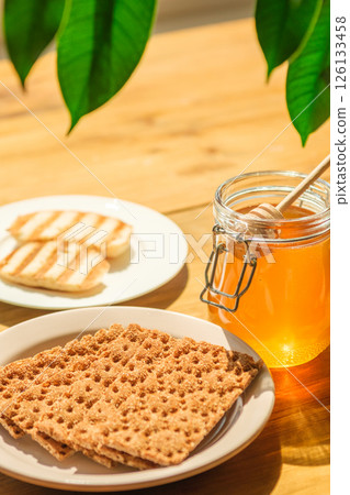 Crispy Crackers and Honey Jar on a Wooden Table, Bathed in Bright Light ideal for Cafes, Bakeries, and Catering Crispy Crackers and Honey Jar on a Wooden Table, Bathed in Bright Light ideal for Cafes, Bakeries, and Catering 126133458