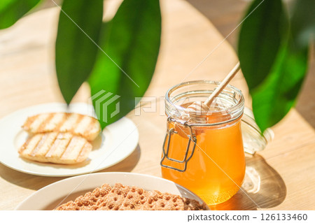 A Jar of Honey With a Wooden Dipper Sits Beside Grilled Bread on a Rustic Table A Jar of Honey With a Wooden Dipper Sits Beside Grilled Bread on a Rustic Table 126133460