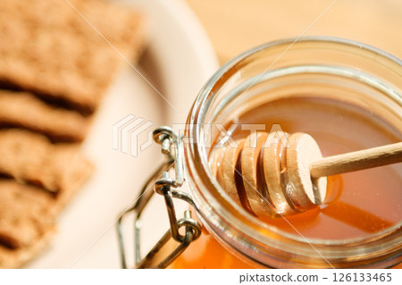 A Glass Jar Filled With Golden Honey Sits With a Wooden Dipper, Accompanied by Crisp Crackers on a Beige Plate in a Cozy Kitchen A Glass Jar Filled With Golden Honey Sits With a Wooden Dipper, Accompanied by Crisp Crackers on a Beige Plate in a Cozy Kitchen 126133465