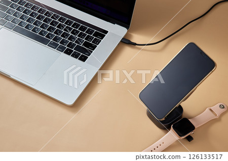 Laptop with smart watch and smartphone charging on wireless charger on beige table, Modern workplace device 126133517