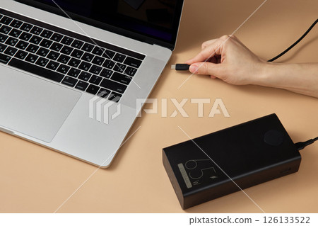 female hand connecting charger cable of power bank to laptop on beige background, close-up. female hand connecting charger cable of power bank to laptop on beige background, close-up. 126133522