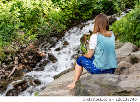 rear view of young woman sitting barefoot on rocks and taking photo of flowing stream with her smartphone Surrounded by lush green foliage, natural setting, walking and hiking 126133740