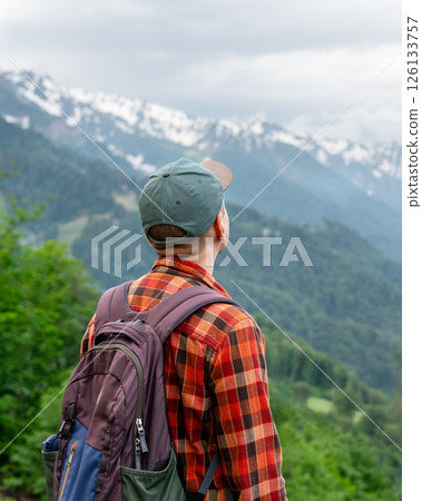 Back view of man wearing in checkered shirt, backpack and cap, looking toward snow-covered peaks, surrounding lush green mountains, hiking and traveling concept Back view of man wearing in checkered shirt, backpack and cap, looking toward snow-covered peaks, surrounding lush green mountains, hiking and traveling concept 126133757