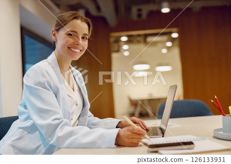 A Smiling Professional Woman Sitting at Her Desk with a Laptop and Notepad in Front of Her 126133931