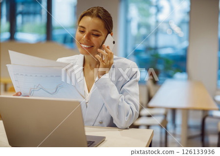 A businesswoman on a call, seated with her laptop and documents in a modern office A businesswoman on a call, seated with her laptop and documents in a modern office 126133936