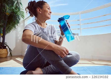 Relaxed Woman Sitting on Yoga Mat Holding a Water Bottle Outdoors 126134102
