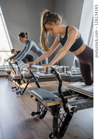 Group of caucasian women exercising with trainer on pilates reformers in a bright studio. Their workout focuses on core strength and flexibility in a modern fitness environment 126134165