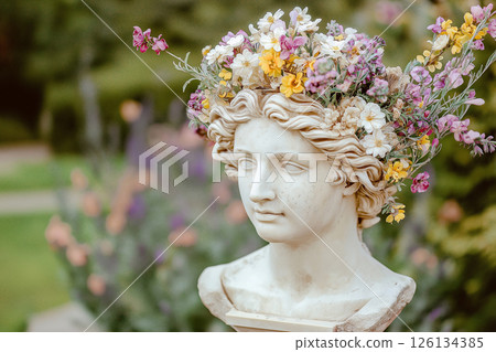 A detailed marble statue head embellished with an arrangement of colorful fresh flowers, set against a soft-focus background of lush greenery, creating a serene and artistic composition. 126134385