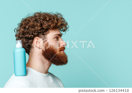 Bearded man with wavy hair holding a blue shampoo bottle on his shoulder 126134418