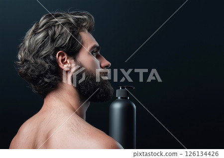 Man with curly hair holding a shampoo bottle against a dark background. Curly haired man with beard with a shampoo bottle standing on his shoulder, highlighting hair care concepts. 126134426