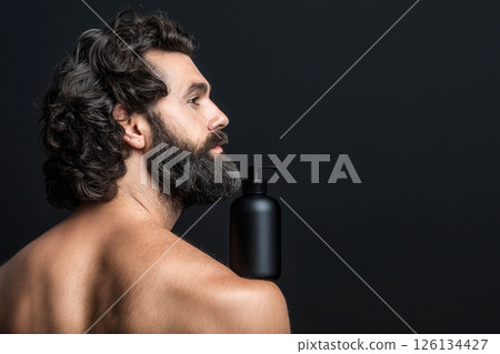 Man with curly hair holding a shampoo bottle against a dark background. Curly haired man with beard with a shampoo bottle standing on his shoulder, highlighting hair care concepts. 126134427