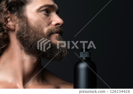 Man with curly hair holding a shampoo bottle against a dark background. Curly haired man with beard with a shampoo bottle standing on his shoulder, highlighting hair care concepts. 126134428