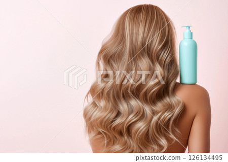 A woman displaying her long, wavy, blonde hair with a focus on a blue shampoo bottle, illustrating clean and healthy hair care enhancing beauty. 126134495
