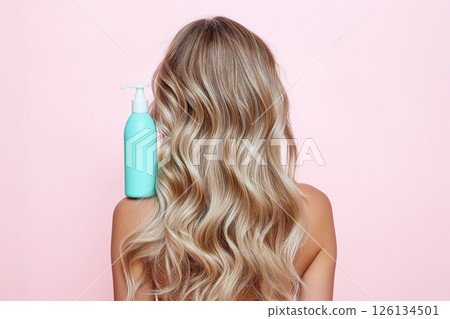 A woman displaying her long, wavy, blonde hair with a focus on a blue shampoo bottle, illustrating clean and healthy hair care enhancing beauty. 126134501