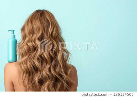 A woman displaying her long, wavy, blonde hair with a focus on a blue shampoo bottle, illustrating clean and healthy hair care enhancing beauty. 126134505