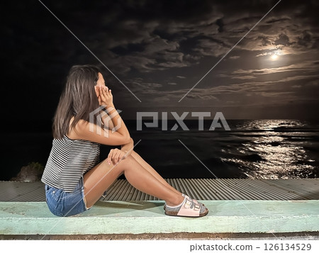 young woman sits at night on the seashore and dreamily looks at the moon in the sky young woman sits at night on the seashore and dreamily looks at the moon in the sky 126134529