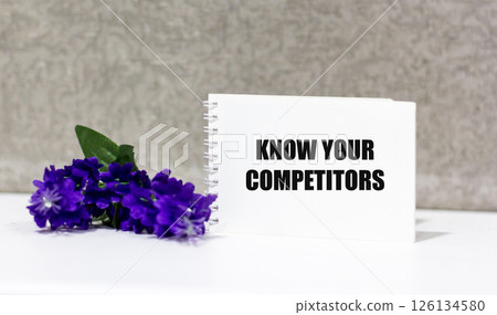 KNOW YOUR COMPETITORS text on a notepad on a gray background next to a blue flower 126134580