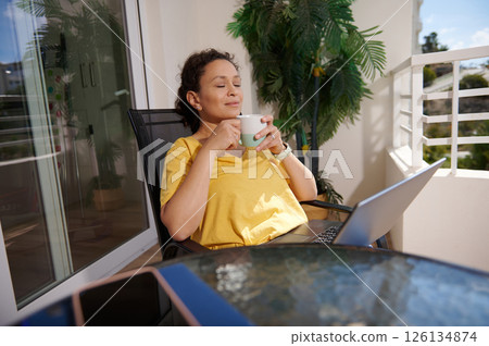 Relaxed Woman Enjoying Coffee Outside While Working Remotely On a Balcony With Laptop 126134874