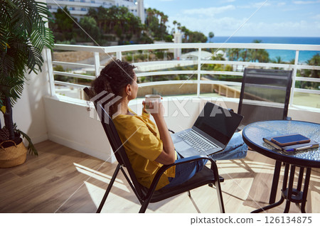 Woman Relaxing on a Balcony with a Laptop and Coffee Overlooking the Ocean Woman Relaxing on a Balcony with a Laptop and Coffee Overlooking the Ocean 126134875