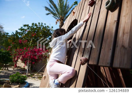 Girl Climbing Outdoor Wooden Wall in Sunny Park with Green Foliage and Blossoms 126134889