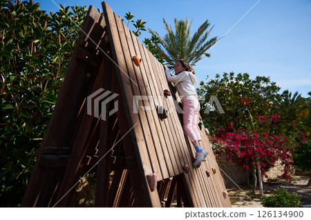 Young Girl Climbing a Wooden Outdoor Play Structure in a Scenic Garden Setting 126134900