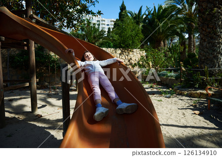 Smiling Child Sliding on Playground Slide Amid Green Palm Trees and Stone Walls Smiling Child Sliding on Playground Slide Amid Green Palm Trees and Stone Walls 126134910