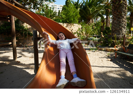 Smiling Young Girl Enjoying a Slide in a Lush Green Playground Smiling Young Girl Enjoying a Slide in a Lush Green Playground 126134911