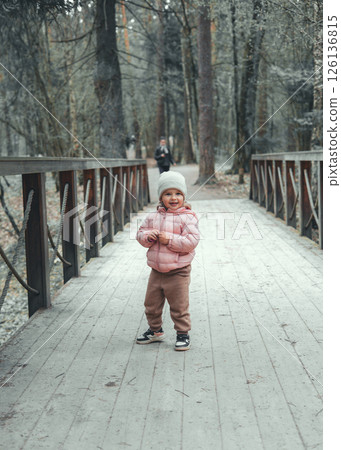 Happy child in pink jacket and hat standing on wooden bridge surrounded by trees in serene winter forest enjoying leisurely walk among beauty of nature. Cute little kid on walk in park. 126136815