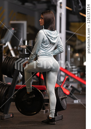 Woman in athletic wear strikes a confident pose in a gym surrounded by weights and fitness equipment. Her focused expression reflects her commitment to health and fitness. 126137154