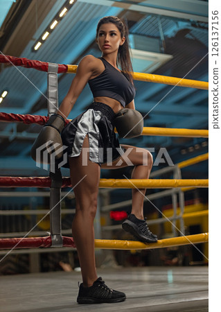 Female boxer stands confidently in a boxing ring wearing training gear. Bright gym lights illuminate her determined expression as she prepares for her workout. Female boxer stands confidently in a boxing ring wearing training gear. Bright gym lights illuminate her determined expression as she prepares for her workout. 126137156