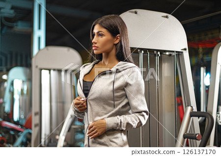 young woman stands in a gym wearing a grey hoodie contemplating her next workout step. The gym is equipped with various exercise machines creating an active atmosphere. 126137170