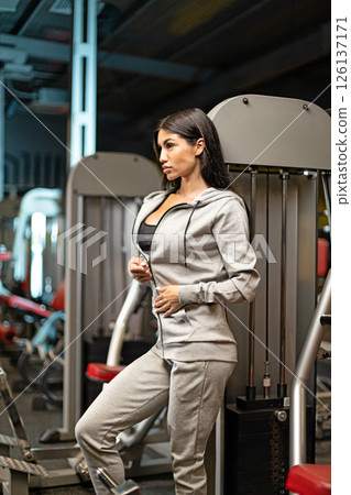 woman in athletic wear rests against gym equipment showcasing her fit physique while taking moment during her workout routine in a well lit fitness center. 126137171