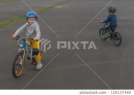 Two children are riding bicycles on an open paved area. One child looks back with a smile, showing joy and confidence while practicing cycling skills together. Two children are riding bicycles on an open paved area. One child looks back with a smile, showing joy and confidence while practicing cycling skills together. 126137340