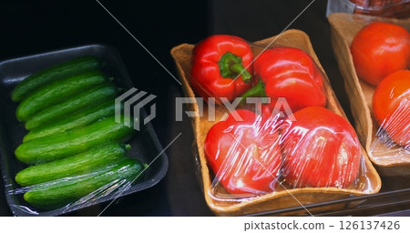 Fresh vegetables including cucumbers, bell peppers, tomatoes, and broccoli gradually appearing, wrapped in plastic film within organized supermarket produce display 126137426