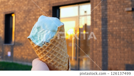 Light blue ice cream in a waffle cone held by a hand in front of a modern brick building with glass doors reflecting a beautiful golden sunset 126137499