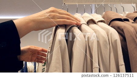 Focused female customer exploring trench coat selection, carefully feeling fabric texture and assessing quality while standing near clothing display rack in retail store interior. Concept fashion 126137511