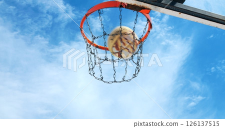 Basketball swishing through metal chain net, arcing against blue clouded sky during outdoor recreational game moment 126137515